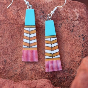 以下が含まれることがあります： 銀色のフックが付いた、ターコイズ、白、オレンジ、ピンクの幾何学模様のイヤリングのペア。 イヤリングは長方形の上部と、山形模様の台形型をしています。 赤茶色の岩の表面に展示されています。