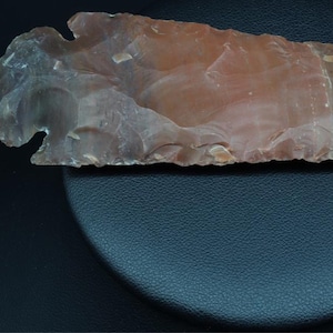 May include: A large, red and gray arrowhead made of stone. The arrowhead is on a black surface.