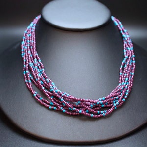 May include: A multi-strand necklace made of small, dark purple and light blue beads. The necklace is layered and creates a cascading effect.