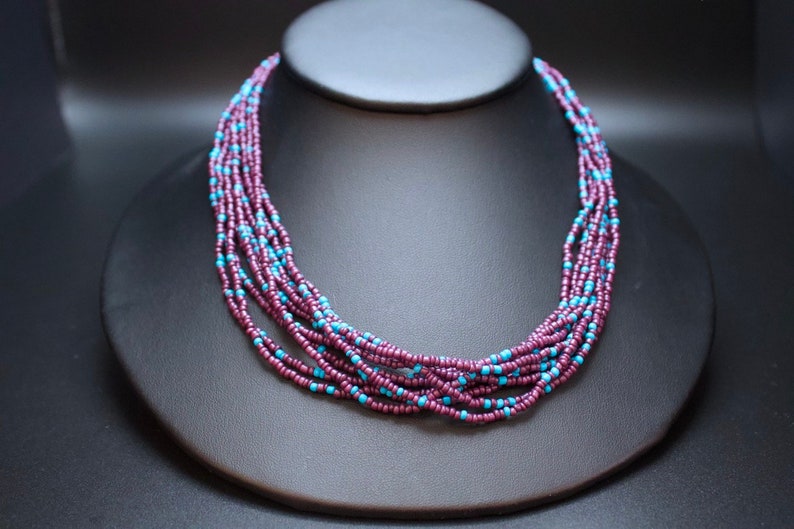 May include: A multi-strand necklace made of small, dark purple and light blue beads. The necklace is layered and creates a cascading effect.
