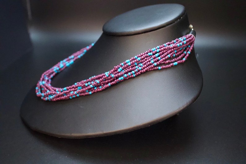 May include: A multi-strand necklace made of small, dark purple and light blue beads. The necklace is displayed on a black mannequin.