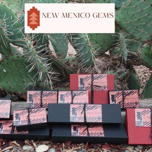 May include: A collection of small, black and red boxes with the text "New Mexico Gems" on them. The boxes are stacked on top of each other and are sitting on a wooden surface.