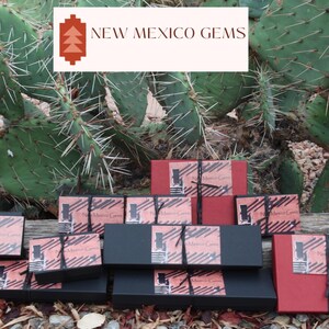 May include: A collection of small, black and red boxes with the text "New Mexico Gems" on them. The boxes are stacked on top of each other and are sitting on a wooden surface.