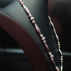 May include: A silver necklace with round beads and elongated links. The necklace is displayed on a black mannequin.