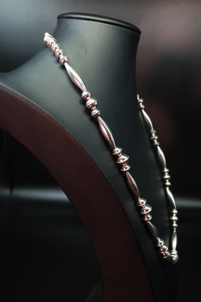 May include: A silver necklace with round beads and elongated links. The necklace is displayed on a black mannequin.