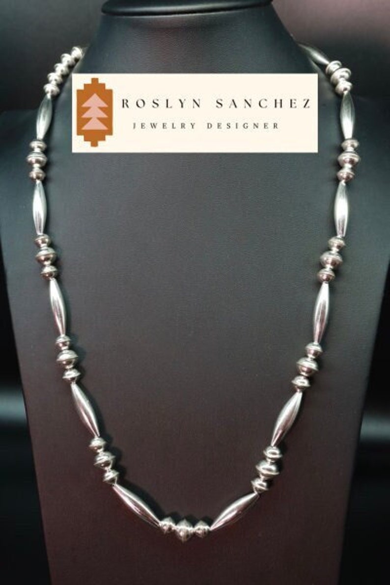 May include: A silver necklace with alternating round and oval beads. The necklace is displayed on a black mannequin bust. The label on the bust reads "Roslyn Sanchez Jewelry Designer".