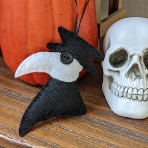 Può includere: Un ornamento di dottore della peste in feltro nero con un becco in feltro bianco e un bottone nero per il naso. L'ornamento è appeso a un nastro nero ed è esposto accanto a un teschio bianco e una zucca arancione.