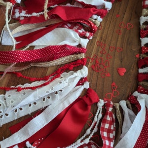 May include: A festive garland featuring red and white fabric strips, ribbons, and lace. The garland includes red polka dots, gingham, and solid red fabrics. Small red heart-shaped confetti is scattered nearby. The garland is tied with twine.