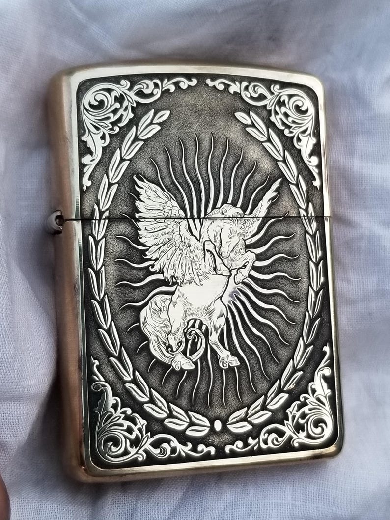 以下が含まれることがあります： 羽根つきの馬が月桂樹の輪で囲まれた彫刻が施された銀色のライター。馬は右を向いており、翼を広げている。背景は黒で、デザインは非常に細かいです。