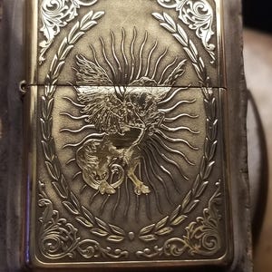 以下が含まれることがあります： 羽根のある生き物が太陽の光を背に、精巧な彫刻が施された金色のかっこいいライター。デザインは月桂樹の輪で囲まれている。
