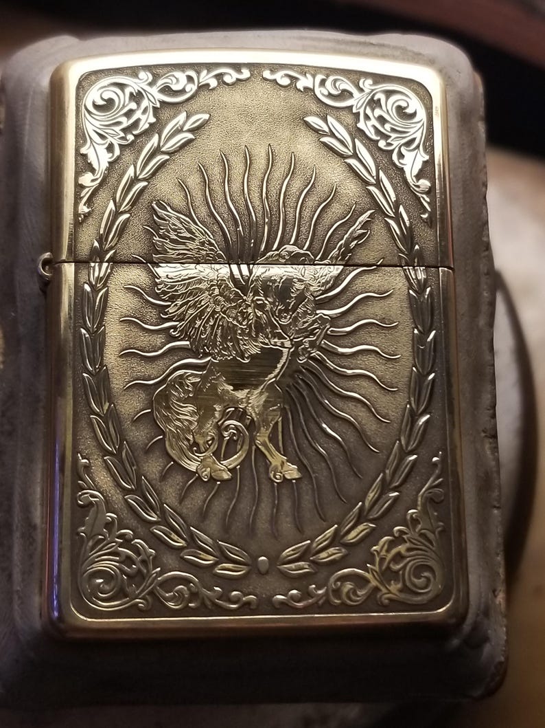 以下が含まれることがあります： 羽根のある生き物が太陽の光を背に、精巧な彫刻が施された金色のかっこいいライター。デザインは月桂樹の輪で囲まれている。