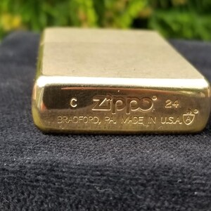 以下が含まれることがあります： 底面に「Zippo」と「Bradford, PA Made in USA」が刻印された金色のジッポーライター。ライターには「24」という数字と「C」という文字が刻印されています。