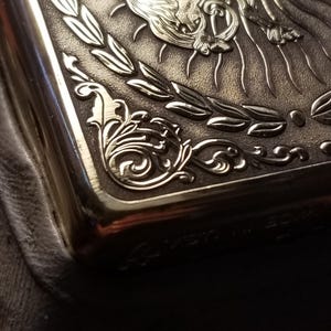 以下が含まれることがあります： 羽根つきの馬と月桂樹の輪のデザインが刻まれた銀色の金属製のタバコケース。ケースの下部には「La  Merveille」という文字が刻まれています。