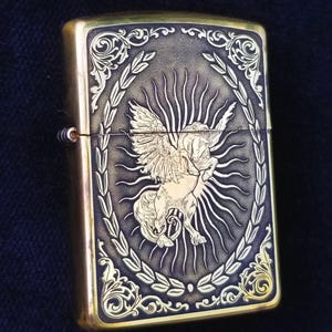 以下が含まれることがあります： 羽根つきの馬が月桂樹の輪で囲まれた、金色の彫刻が施されたライター。馬は右を向いており、背後には太陽の光が輝いています。