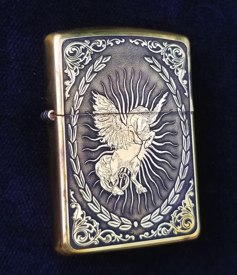 以下が含まれることがあります： 羽根つきの馬が月桂樹の輪で囲まれた、金色の彫刻が施されたライター。馬は右を向いており、背後には太陽の光が輝いています。