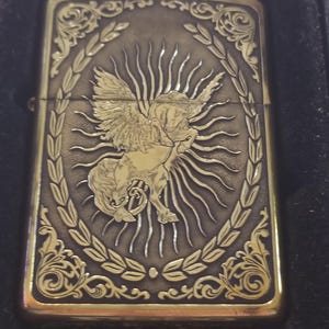以下が含まれることがあります： 金色に輝くジッポーライター。羽根つきの馬が太陽の光の中に描かれ、その周りを月桂樹の輪が囲んでいます。ライターの上部には「Armor Case」と「Made in USA」の文字が刻まれています。
