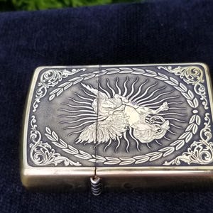 以下が含まれることがあります： 羽根のある生き物が太陽の光を背にしている彫刻が施された金色のかっこいいライター。ライターは、細かい花柄の縁取りがされている。
