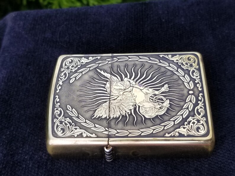 以下が含まれることがあります： 羽根のある生き物が太陽の光を背にしている彫刻が施された金色のかっこいいライター。ライターは、細かい花柄の縁取りがされている。