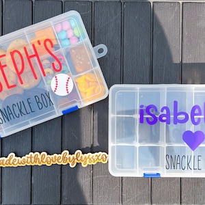 May include: Two clear plastic snack boxes. One is filled with snacks and labeled "Joseph's Snack Box" with a baseball sticker. The other is empty and labeled "Isabella's Snack Box" with a purple heart.