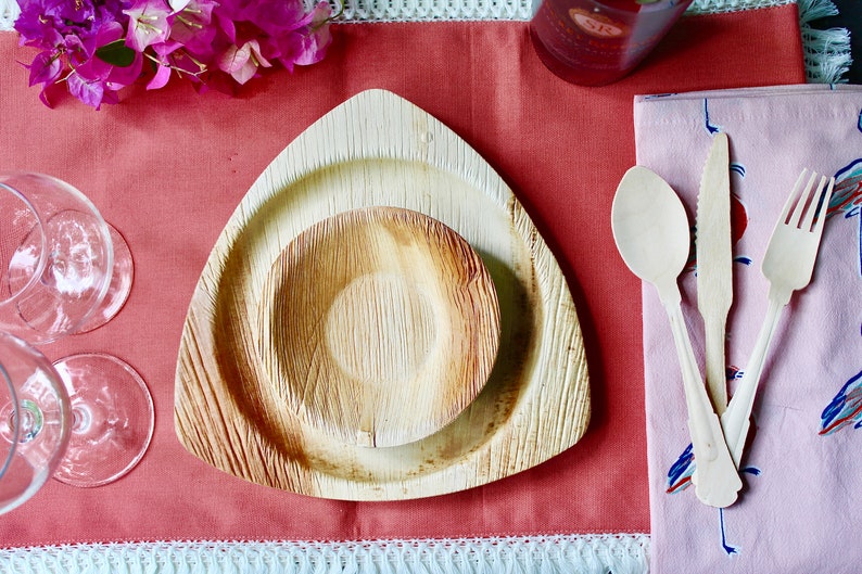 May include: A set of two triangular palm leaf plates, one smaller than the other, on a red tablecloth. The plates are made of natural materials and are perfect for eco-friendly dining.
