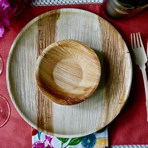 May include: A table setting with a large, round, brown and white palm leaf plate, a smaller brown palm leaf bowl, and wooden cutlery on a red tablecloth.