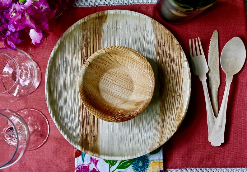 May include: A table setting with a large, round, brown and white palm leaf plate, a smaller brown palm leaf bowl, and wooden cutlery on a red tablecloth.