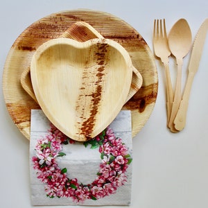 May include: Two brown palm leaf plates, one shaped like a heart, with a floral patterned napkin on top.  A set of three wooden utensils, including a fork, knife, and spoon, are placed to the right of the plates.