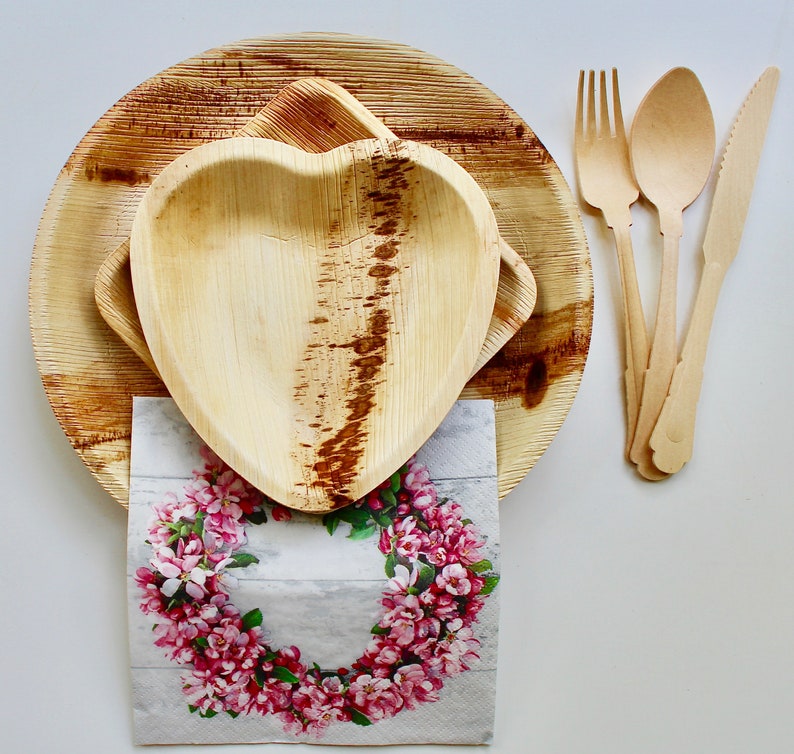 May include: Two brown palm leaf plates, one shaped like a heart, with a floral patterned napkin on top.  A set of three wooden utensils, including a fork, knife, and spoon, are placed to the right of the plates.