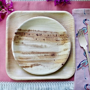 May include: A set of two round, brown, palm leaf plates with a square, brown, palm leaf plate. The plates are set on a pink table runner with a white fringe. A pink napkin with a flamingo print is on the right side of the image. The napkin has a wooden fork, knife, and spoon on top of it.