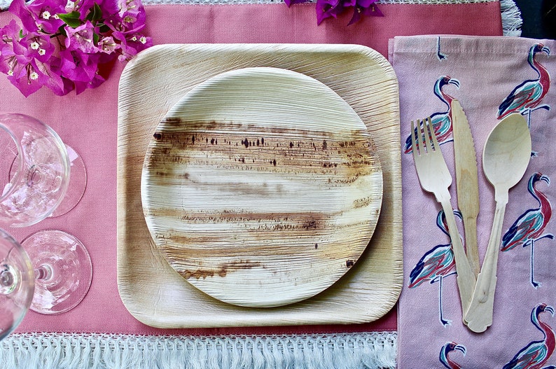 May include: A set of two round, brown, palm leaf plates with a square, brown, palm leaf plate. The plates are set on a pink table runner with a white fringe. A pink napkin with a flamingo print is on the right side of the image. The napkin has a wooden fork, knife, and spoon on top of it.