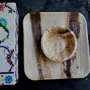 May include: A square, light brown, palm leaf plate with a smaller, round, palm leaf bowl in the center. Both are made from natural materials and have a rustic, organic look.