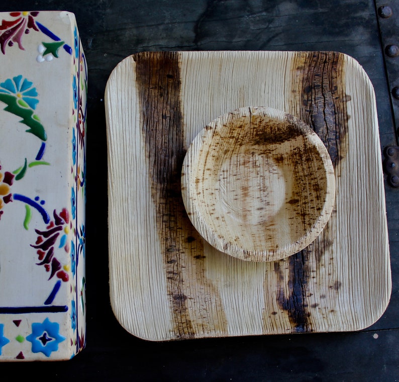 May include: A square, light brown, palm leaf plate with a smaller, round, palm leaf bowl in the center. Both are made from natural materials and have a rustic, organic look.