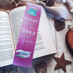 Pode incluir: Um marcador de página rosa e roxo com textura de madeira. Apresenta uma chávena de chá, uma pilha de livros e o texto "Drink good tea Read good books."