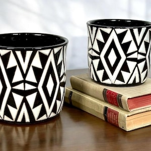 May include: Two black and white patterned ceramic pots with geometric designs. The pots are cylindrical with a black interior and sit on top of two vintage books with red and tan covers. The pots are decorative.