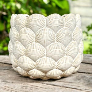 May include: Cream-colored ceramic planter with a seashell pattern. The planter has a round shape and is covered in overlapping, fan-shaped seashell designs. The surface has a textured appearance, and the planter sits on a wooden surface.