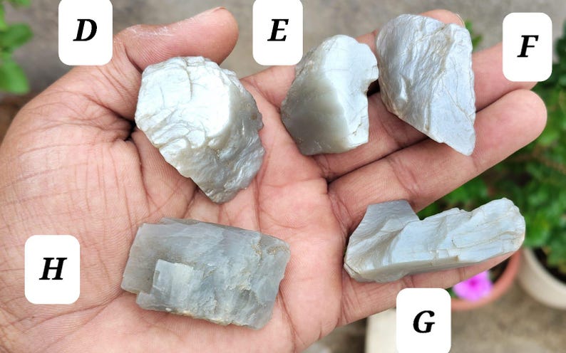 May include: Five raw, translucent, light gray moonstone specimens labeled D, E, F, G, and H. Each stone features an irregular, jagged shape with a smooth, reflective surface texture and visible internal crystalline layers and fractures.