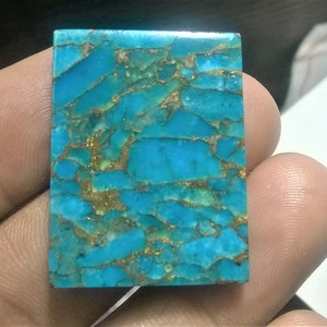 May include: A rectangular turquoise cabochon with gold pyrite inclusions. The stone has a bright blue color and a smooth, polished surface.