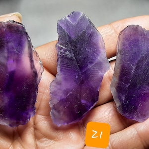 K&ouml;nnte beinhalten: Drei rohe Amethyst-Kristallscheiben in verschiedenen Formen und Gr&ouml;&szlig;en, die tiefe lilafarbene Farbt&ouml;ne mit helleren, durchscheinenden Bereichen aufweisen. Die rauen Kanten und nat&uuml;rlichen Texturen der Steine sind sichtbar. Die Kristalle werden in einer Hand gehalten.