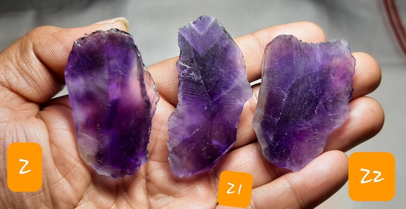 K&ouml;nnte beinhalten: Drei rohe Amethyst-Kristallscheiben in verschiedenen Formen und Gr&ouml;&szlig;en, die tiefe lilafarbene Farbt&ouml;ne mit helleren, durchscheinenden Bereichen aufweisen. Die rauen Kanten und nat&uuml;rlichen Texturen der Steine sind sichtbar. Die Kristalle werden in einer Hand gehalten.