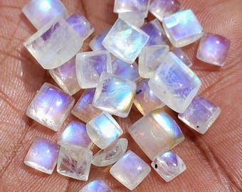 Könnte beinhalten: Eine Ansammlung kleiner, quadratisch geschliffener Mondstein-Edelsteine mit einer durchscheinenden weißen Basis und irisierenden blauen sowie violetten Lichtreflexen. Jeder Stein hat eine glatte, polierte Oberfläche mit sichtbaren Einschlüssen.
