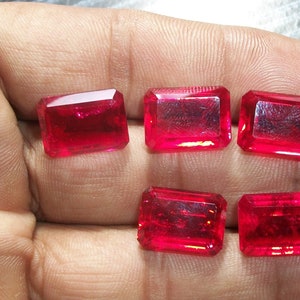 May include: Six red gemstone rectangular cut cabochons, likely rubies, displayed on a hand.