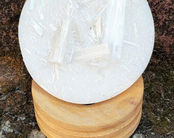 May include: A circular white translucent resin disc embedded with small selenite crystal shards, resting atop a cylindrical light brown wooden base. The resin surface features a textured, speckled appearance with three prominent crystal pieces.
