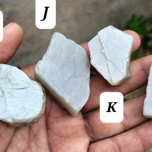 May include: Four irregular, pale gray, semi-translucent stones labeled I, J, K, and L. Each stone features a smooth, matte surface with subtle, fine, vein-like cracks running across the exterior, showcasing a natural, unpolished, and raw appearance.