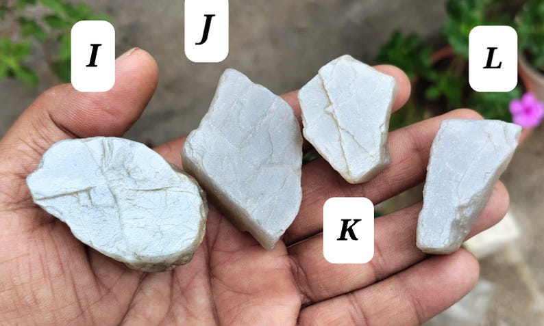 May include: Four irregular, pale gray, semi-translucent stones labeled I, J, K, and L. Each stone features a smooth, matte surface with subtle, fine, vein-like cracks running across the exterior, showcasing a natural, unpolished, and raw appearance.