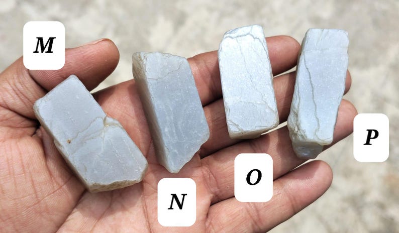 May include: Four rough, pale blue-grey stones labeled M, N, O, and P. Each stone has an irregular, angular shape with visible surface textures, natural cracks, and a matte finish. The stones are presented in a row, showcasing their distinct forms.