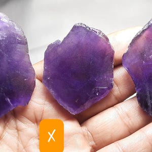 K&ouml;nnte beinhalten: Drei rohe Amethystkristalle in verschiedenen Formen und Gr&ouml;&szlig;en, die einen tiefvioletten Farbton mit helleren Bereichen aufweisen. Die Kristalle werden in einer Hand gehalten und zeigen ihre nat&uuml;rlichen, rauen Texturen. Das Bild ist gut beleuchtet.