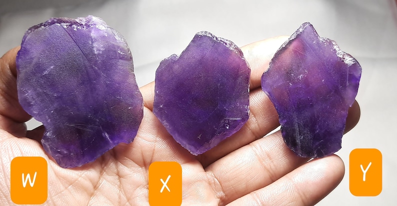K&ouml;nnte beinhalten: Drei rohe Amethystkristalle in verschiedenen Formen und Gr&ouml;&szlig;en, die einen tiefvioletten Farbton mit helleren Bereichen aufweisen. Die Kristalle werden in einer Hand gehalten und zeigen ihre nat&uuml;rlichen, rauen Texturen. Das Bild ist gut beleuchtet.