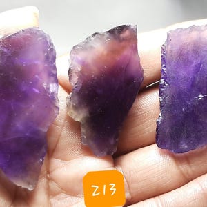 K&ouml;nnte beinhalten: Drei rohe Amethystkristallproben in Lila- und Lavendelt&ouml;nen. Jeder Stein hat eine einzigartige Form und Textur mit sichtbaren kristallinen Strukturen. Die Kristalle werden in einer Hand gehalten, mit orangefarbenen Etiketten mit der Aufschrift "Z12", "Z13" und "Z14".