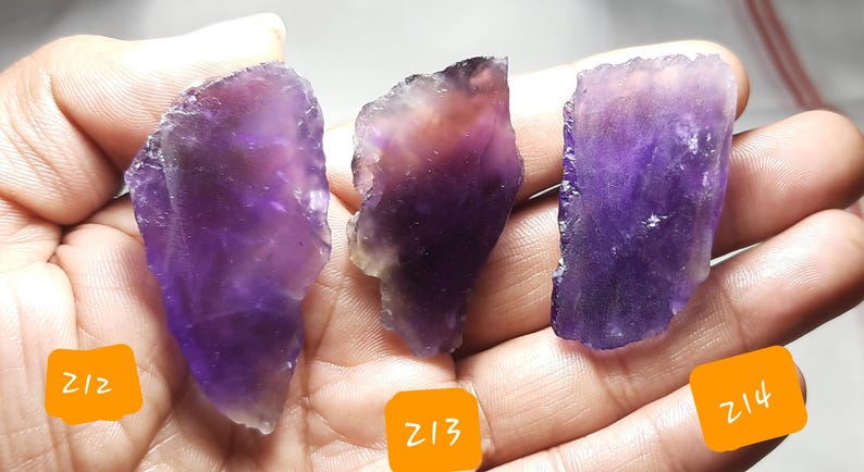 K&ouml;nnte beinhalten: Drei rohe Amethystkristallproben in Lila- und Lavendelt&ouml;nen. Jeder Stein hat eine einzigartige Form und Textur mit sichtbaren kristallinen Strukturen. Die Kristalle werden in einer Hand gehalten, mit orangefarbenen Etiketten mit der Aufschrift "Z12", "Z13" und "Z14".