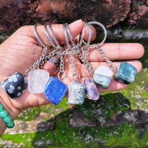 May include: A collection of seven keychains featuring polished, tumbled gemstones in various colors including black with white spots, clear, blue, reddish-brown, mottled green, purple, and a dark green with white inclusions, all attached to silver metal rings.